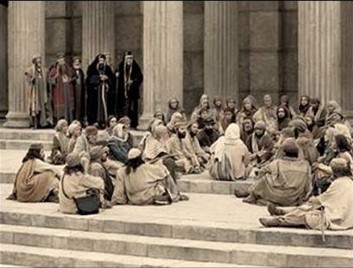 A crowd of humbly dressed people sit in a circle on the floor of a temple around someone with their head covered. They look relaxed. Religious figures are seen standing afar off. 