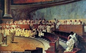 A large group of seated men in white togas surround a stage where someone is speaking. It appears to be an indoor amphitheater. 
