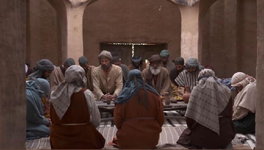 Around twenty people sit around a table with simple food in an ancient dwelling bowing their heads and folding their hands.